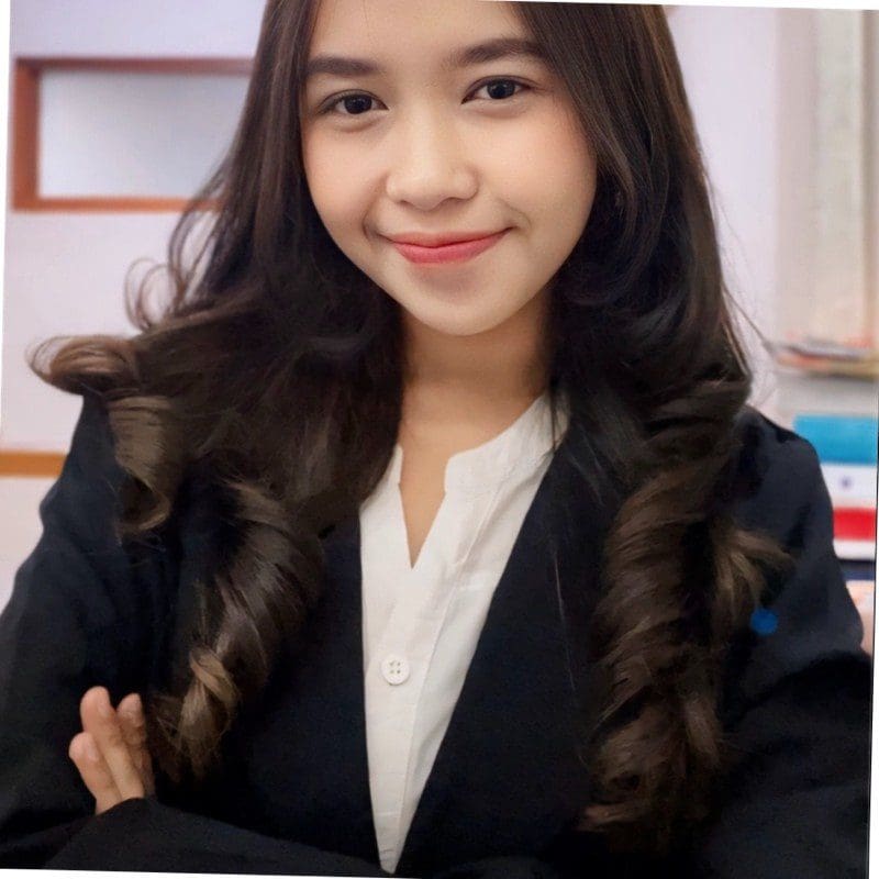 Seorang wanita muda berambut kerinting panjang tersenyum dalam pakaian profesional, dengan blazer hitam dan kemeja putih, bergambar dengan tangan disilangkan.
