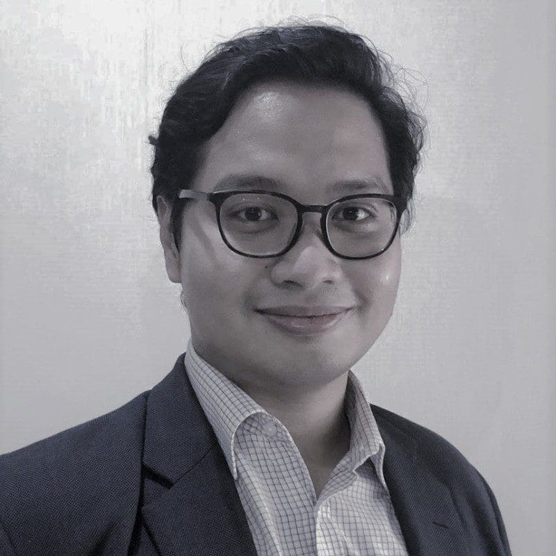 Seorang individu yang kelihatan profesional memakai cermin mata dan sut, tersenyum dengan latar belakang neutral.