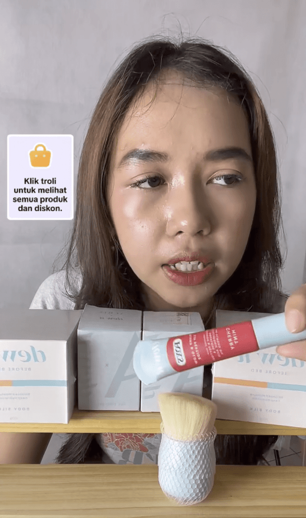 Primer plano de una mujer joven mostrando un producto cosmético durante una retransmisión en directo, con varios productos en cajas detrás de ella.