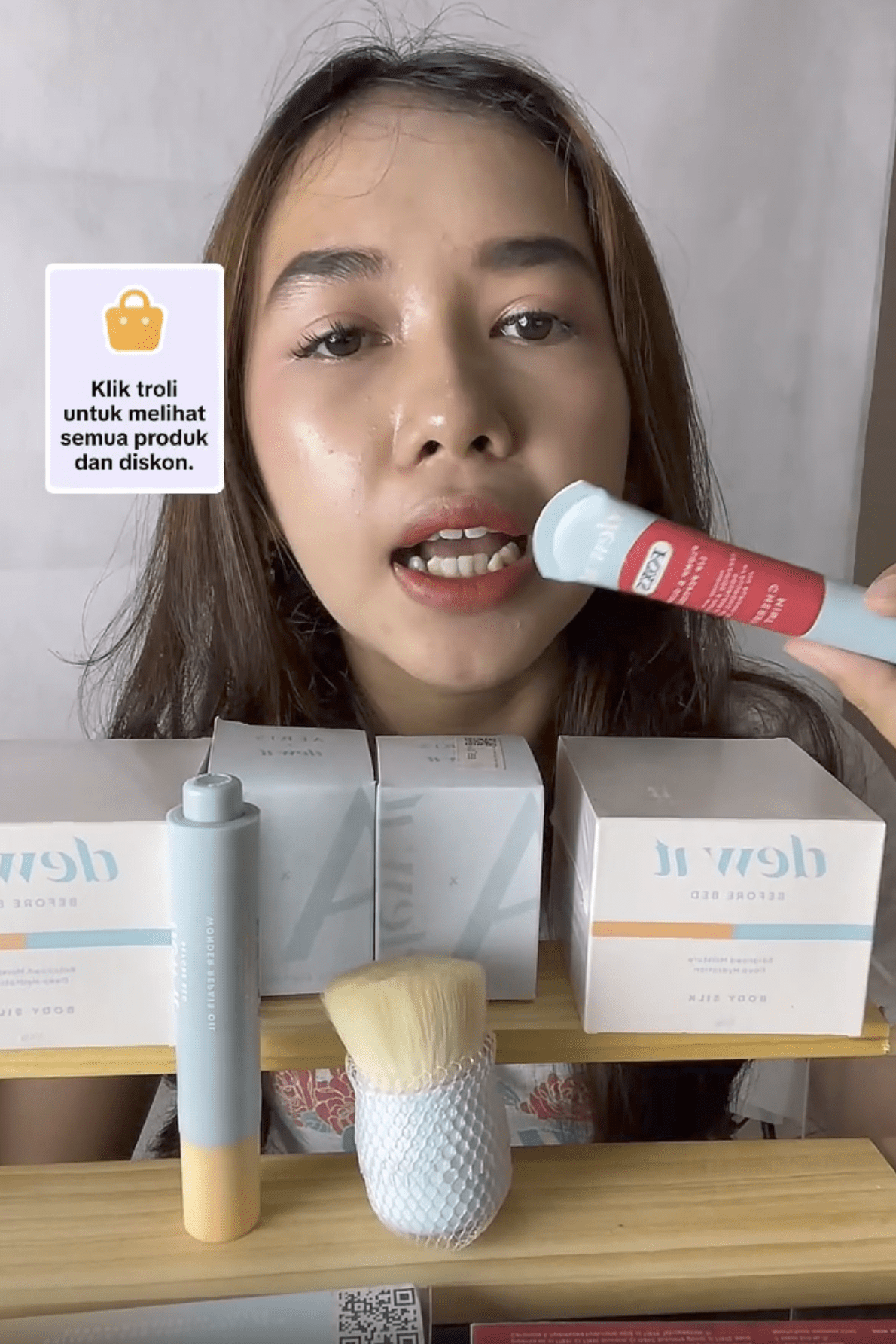 Seorang wanita muda memegang produk penjagaan kulit sambil mempromosikannya semasa strim langsung, dengan kotak pelbagai produk dipaparkan di belakangnya.