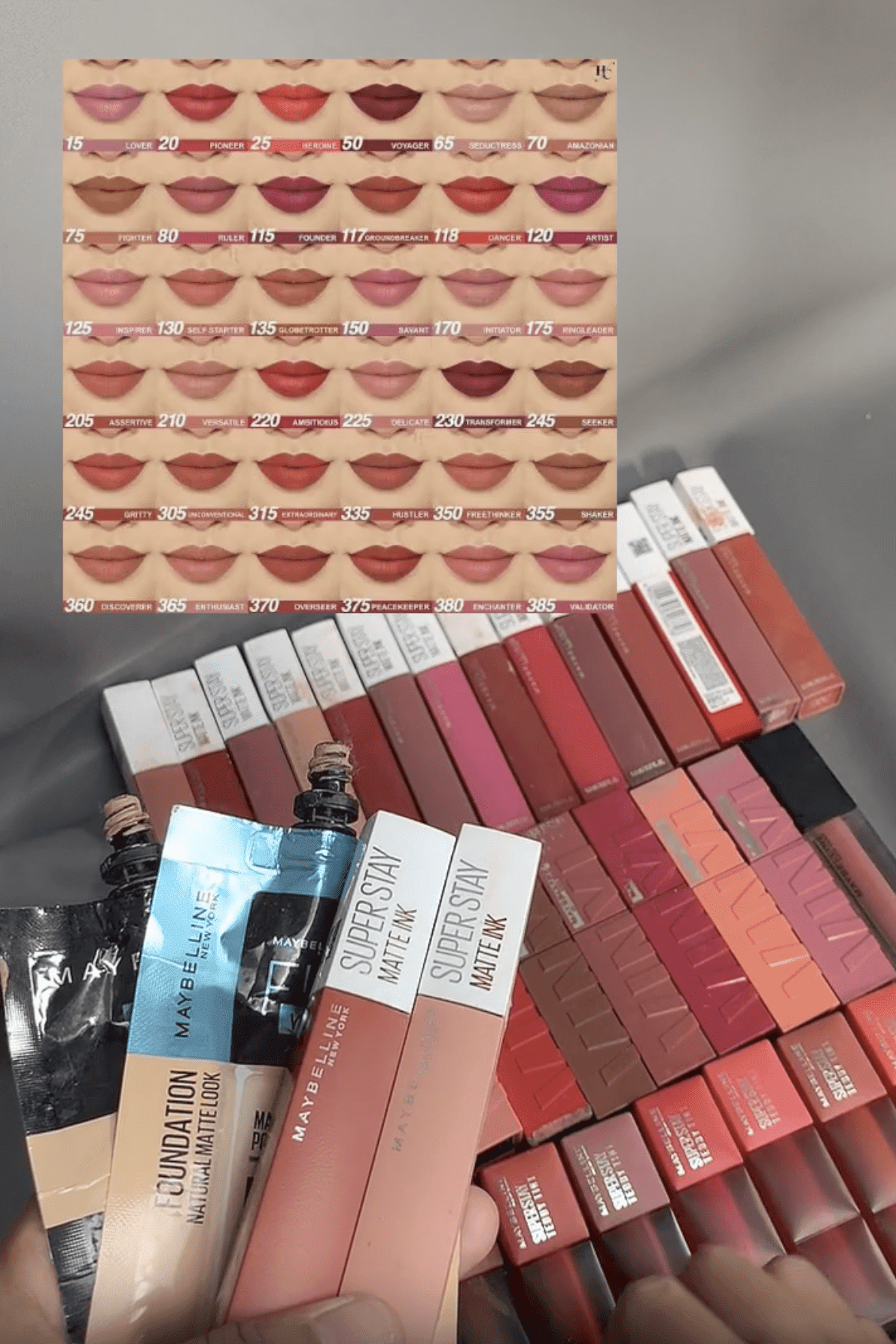 Imej menunjukkan carta swatch warna pelbagai rona bibir dengan nombor yang sepadan, bersama beberapa produk solek Maybelline termasuk bedak asas dan gincu cecair yang disusun rapi.