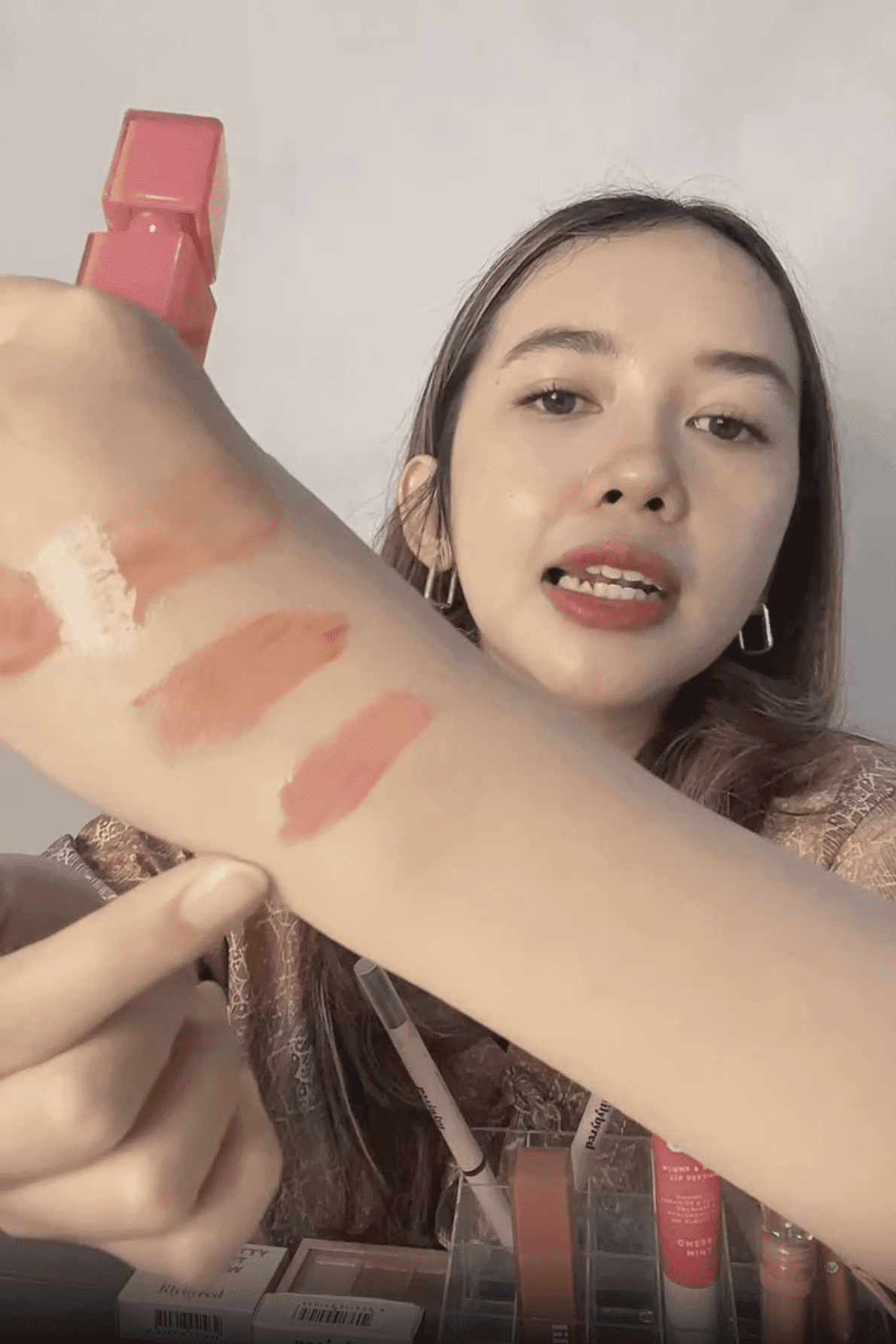Seorang wanita muda mempamerkan swatch produk bibir pada lengannya semasa strim langsung, dengan pelbagai warna bibir di latar belakang.