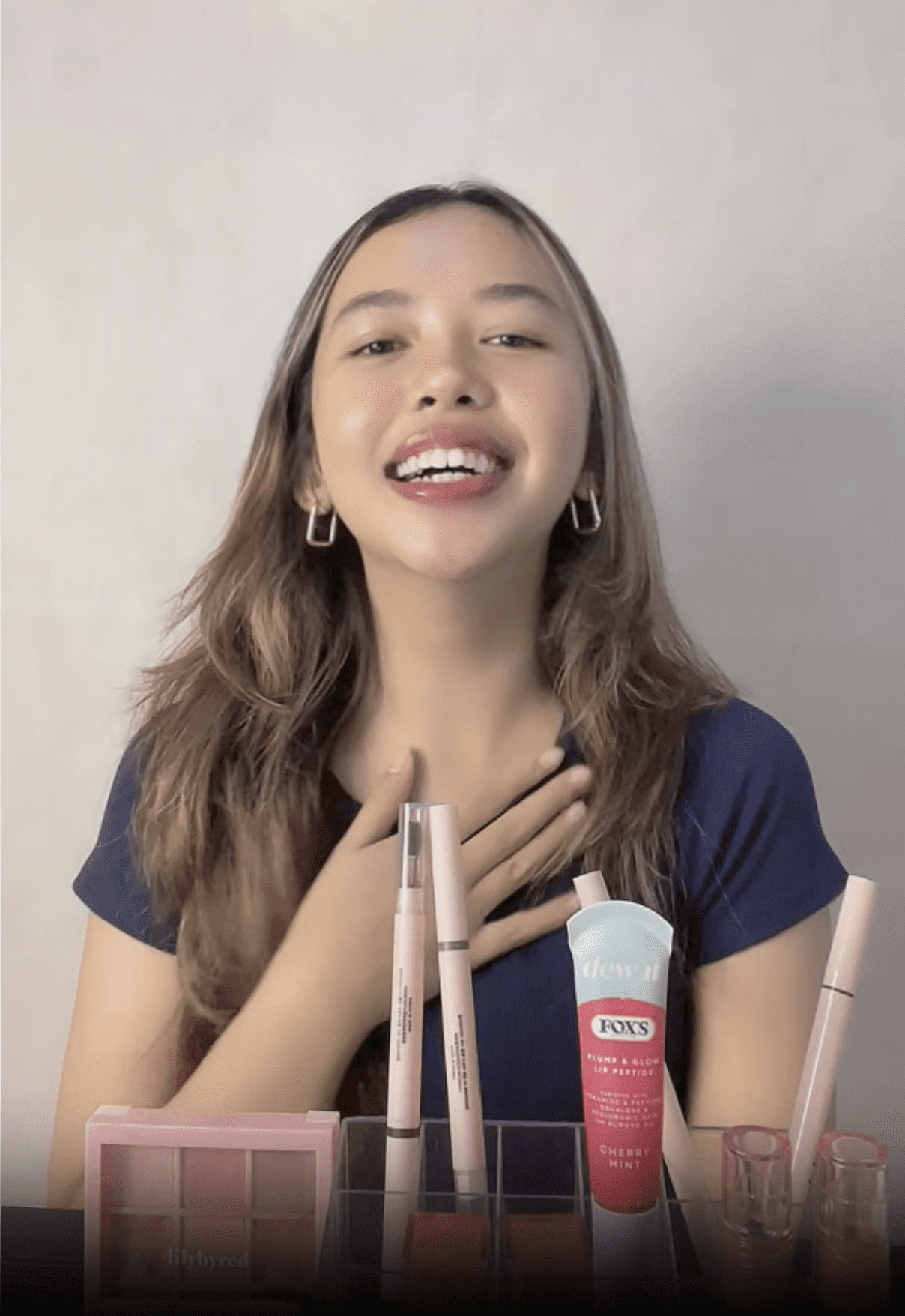 Hos strim langsung wanita yang tersenyum dengan baju biru mempamerkan produk kecantikan di atas meja, berinteraksi dengan penonton.
